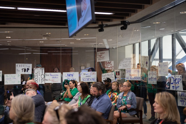 Members of AFSCME Local 34 and Local 2822 hold signs against a glass partition separating the County board chambers.
